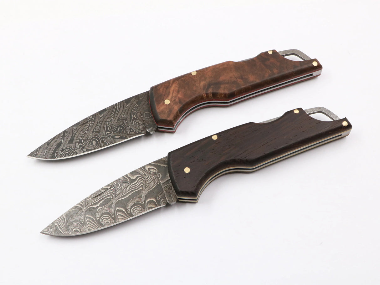 Zwei Waldflamme Taschenmesser nebeneinander: links mit dunklem Wengeholzgriff, rechts mit warmem Walnussholz, beide mit Damastklinge auf Naturhintergrund