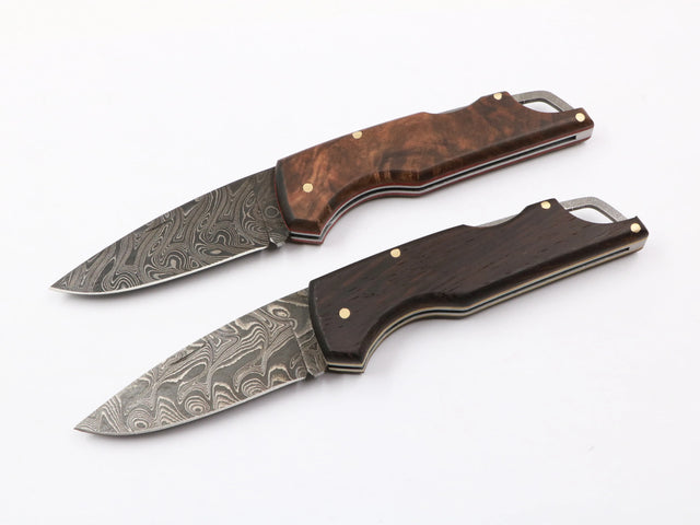 Zwei Waldflamme Taschenmesser nebeneinander: links mit dunklem Wengeholzgriff, rechts mit warmem Walnussholz, beide mit Damastklinge auf Naturhintergrund